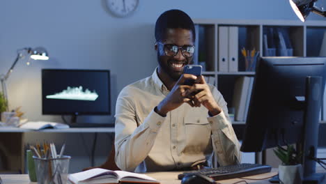 Cheerful-Young-Man-Texting-A-Message-On-The-Smartphone-And-Smiling-In-The-Office-At-Night