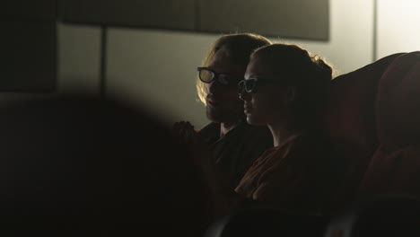 Side-View-Of-Couple-Wearing-3D-Glasses-Sitting-In-The-Cinema-While-They-Watching-A-Movie-And-Eating-Popcorn-With-The-Lights-Off