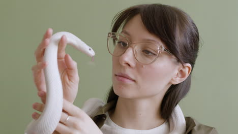 Portrait-Of-A-Young-Woman-With-Eyeglasses-Holding-A-White-Pet-Snake-Around-Her-Neck-And-Hands-On-A-Green-Background-2