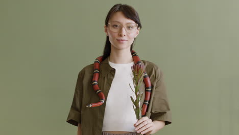 Portrait-Of-A-Young-Woman-With-A-Pet-Snake-Around-Her-Neck-Holding-A-Twig-And-Looking-At-The-Camera-On-A-Green-Background