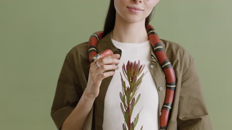 Portrait-Of-A-Young-Woman-With-A-Pet-Snake-Around-Her-Neck-Holding-A-Twig-On-A-Green-Background