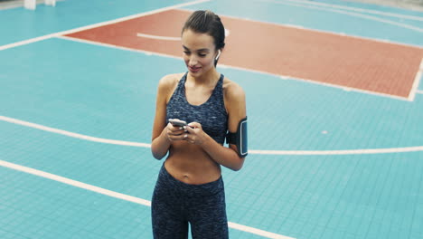 Sporty-Girl-With-Airpods-Texting-Message-On-Smartphone-While-Standing-At-Outdoor-Court-On-A-Summer-Day