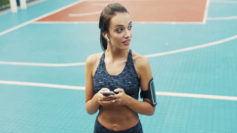 Sportliches-Mädchen-Mit-Airpods-SMS-Nachricht-Auf-Dem-Smartphone,-Während-Es-An-Einem-Sommertag-Auf-Dem-Außenplatz-Steht-1