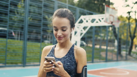 Sporty-Girl-With-Airpods-Laughing-While-Texting-Message-On-Smartphone-At-Outdoor-Court-On-A-Summer-Day