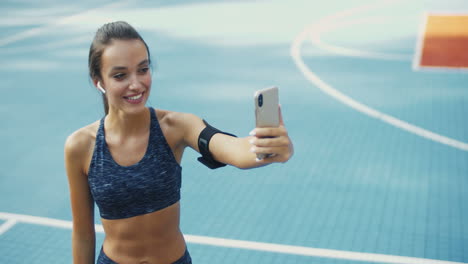 Cheerful-Sporty-Woman-With-Airpods-Smiling-And-Showing-Biceps-To-Smartphone-Camera-While-Having-Videochat-At-Sport-Court