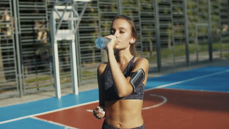 Sportliche-Frau,-Die-An-Einem-Außenplatz-Steht,-Sich-Ausruht-Und-Nach-Dem-Training-Kaltes-Wasser-Trinkt