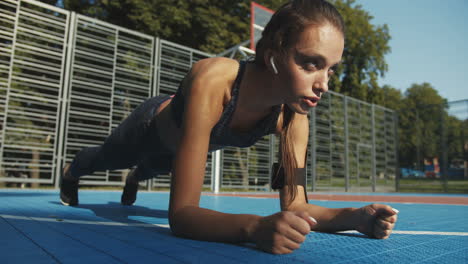 Focused-Fitness-Girl-With-Airpods-Doing-Plank-Exercise-And-Breathing-Deep-At-Sport-Court-On-Summer-Day