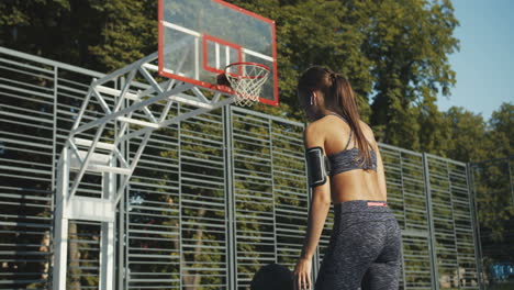 Rückansicht-Einer-Sportlichen-Frau-Mit-Airpods,-Die-An-Einem-Sonnigen-Tag-Auf-Dem-Basketballplatz-Mit-Ball-Spielt