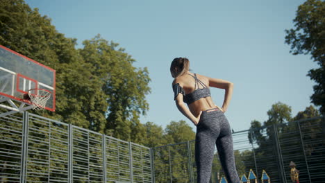 Rear-View-Of-A-Sporty-Woman-Warming-Up-And-Stretching-At-Outdoor-Basketball-Court-On-A-Summer-Morning