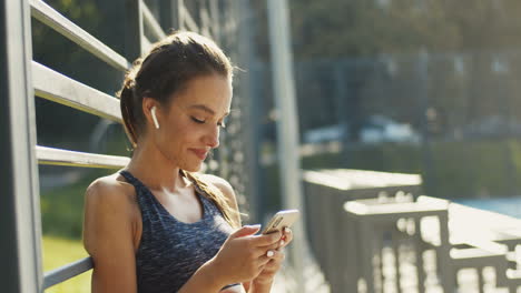 Sporty-Girl-With-Airpods-Laughing-While-Texting-Message-On-Smartphone-After-Workout-At-Outdoor-Court-On-A-Summer-Day