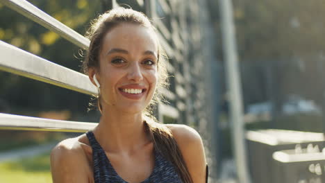 Portrait-Of-A-Cheerful-Sporty-Woman-With-Airpods-Smiling-At-The-Camera-After-Workout-At-Outdoor-Court-On-Sunny-Day