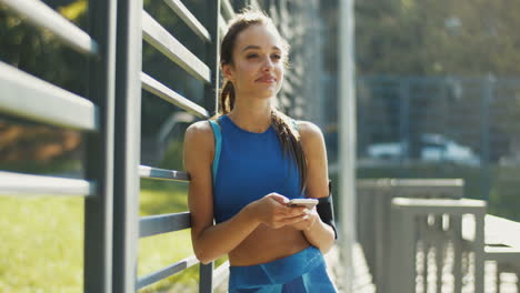 Pretty-Sportswoman-Texting-Message-On-Smartphone-And-Smiling-At-Outdoor-Court-On-A-Summer-Day