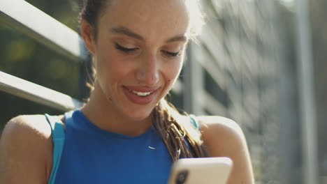 Close-Up-Of-A-Pretty-Sportswoman-Texting-Message-On-Smartphone-And-Smiling-At-Outdoor-Court-On-A-Summer-Day