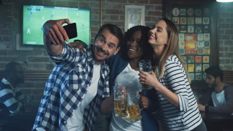Young-Multiethnic-Friends-In-The-Pub-During-Sport-Game,-Man-Taking-Selfie-Photo-On-The-Smartphone-With-Two-Women
