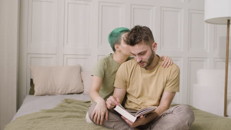 Young-Man-Sitting-On-The-Bed-And-Leafing-Through-A-Book-While-His-Girlfriend-Cuddling-Him
