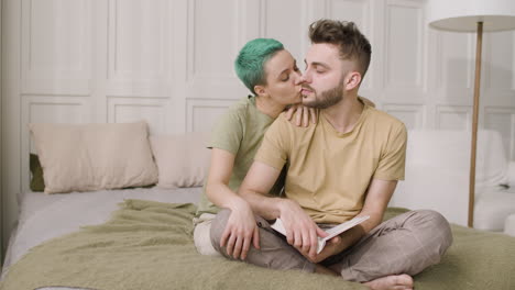 Young-Man-Sitting-On-The-Bed-And-Holding-A-Book-While-His-Girlfriend-Kissing-And-Embracing-Him