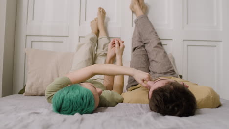 Loving-Couple-Holding-Hands-And-Looking-At-Each-Other-While-Lying-On-A-Cozy-Bed-With-Their-Legs-Up-On-The-Wall