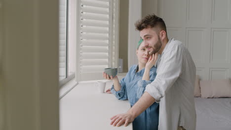 Young-Woman-Next-To-The-Window-Holding-A-Mug-And-Gently-Caressing-Her-Boyfriend-Who-Standing-Behind-Her