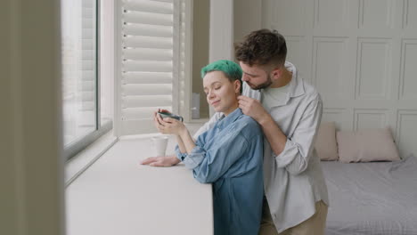 Young-Man-Tenderly-Cuddling-His-Girlfriend-Who-Holding-A-Mug-While-Standing-Together-Next-To-The-Window-In-The-Bedroom