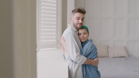Loving-Couple-Tenderly-Hugging-And-Looking-At-The-Camera-While-Standing-Next-To-The-Window-In-The-Bedroom