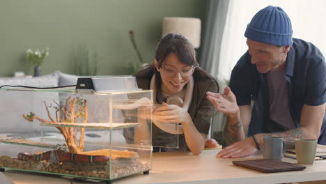 Woman-Holding-A-White-Snake-And-Talking-With-Her-Boyfriend-Leaning-On-A-Table-With-Terrarium