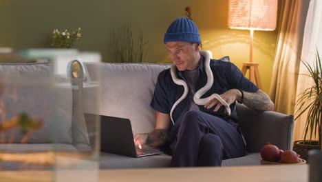 Man-Sitting-On-A-Sofa-And-Holding-A-Snake-Around-His-Neck-While-Using-A-Laptop-At-Home