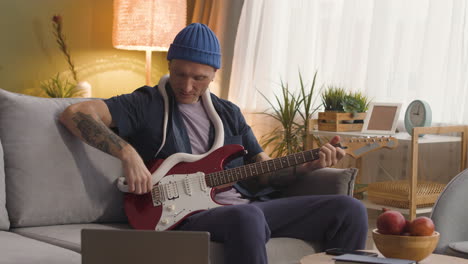 Man-Sitting-On-A-Sofa-And-Holding-A-Snake-Around-His-Neck-While-Playing-The-Electric-Guitar
