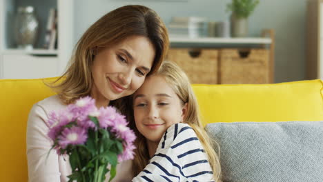 Clore-Up-View-Of-Mother-Hugging-Her-Cute-Teenager-Daughter-Sitting-On-Sofa-While-Holding-A-Bouquet-Of-Flowers