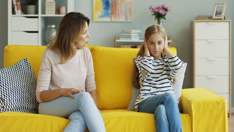 Blonde-Mother-Sitting-With-Her-Teenage-Daughter-On-Yellow-Sofa-And-Quarreling-At-Her,-Girl-Doesn'T-Listen-And-Covers-Her-Ears