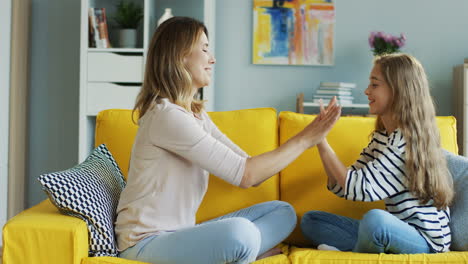 Blonde-Mother-With-Her-Cute-Daughter-Playing-Hands-Game-While-Resting-On-Yellow-Sofa-In-Living-Room