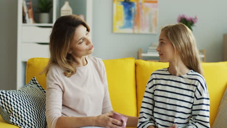 Blonde-Mother-Holding-A-Cup-Of-Coffe-And-Sitting-With-Her-Teenage-Daughter-On-Yellow-Couch-At-Home-And-Having-A-Nice-Talk