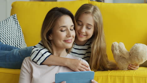 Blonde-Mother-And-Daughter-Resting-On-The-Sofa-At-Home-And-Watching-Something-On-A-Tablet