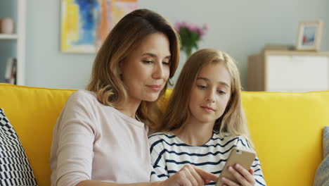 Cute-Little-Girl-With-Her-Mother-Scrolling-On-The-Smartphone-And-Talking-While-Sitting-On-Sofa-In-Living-Room
