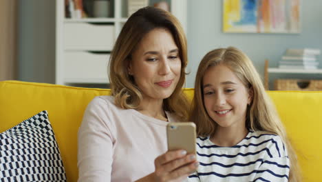 Blonde-Mother-With-Her-Teen-Daughter-Watching-Video-On-Smartphone,-Talking-And-Smiling-While-They-Are-Sitting-On-Sofa-In-Living-Room
