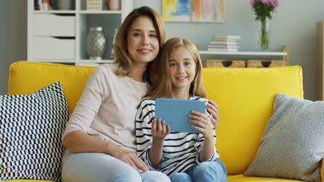 Blonde-Mother-With-Her-Teen-Daughter-Watching-Video-On-Tablet,-Talking-And-Smiling-While-They-Are-Sitting-On-Sofa-In-Living-Room