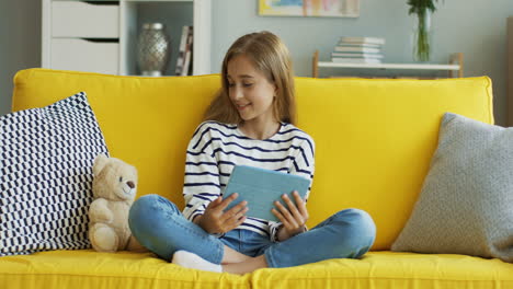 Blonde-Teenage-Girl-Sitting-On-Yellow-Sofa-At-Home-And-Watching-Something-On-Tablet,-Then-She-Looks-And-Smiles-At-Camera