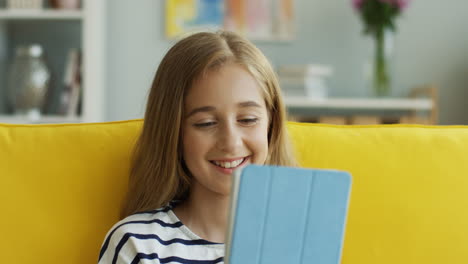 Close-Up-View-Of-Blonde-Teenage-Girl-Sitting-On-Yellow-Sofa-At-Home-And-Watching-Something-On-Tablet