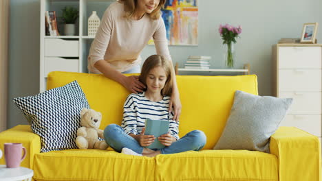 Blonde-Cute-Girl-Sitting-On-Yellow-Couch-At-Home-And-Watching-Something-On-Tablet,-Then-Her-Mother-Approachs-And-Hugs-Her-1