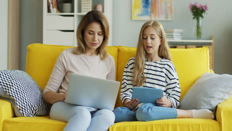 Blonde-Mutter-Und-Tochter-Sitzen-Auf-Einer-Gelben-Couch,-Während-Sie-Sich-Unterhalten-Und-Laptop-Und-Tablet-Im-Wohnzimmer-Benutzen