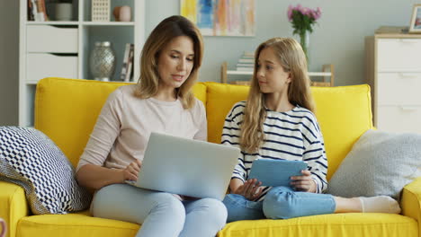Blonde-Mother-And-Daughter-Sitting-On-Yellow-Couch-While-They-Talking-And-Using-Laptop-Computer-And-Tablet-In-The-Living-Room-1