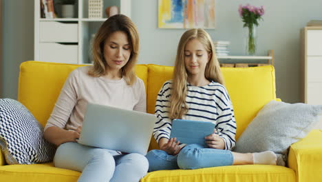 Blonde-Mother-And-Daughter-Sitting-On-Yellow-Couch-While-They-Talking-And-Using-Laptop-Computer-And-Tablet-In-The-Living-Room-2