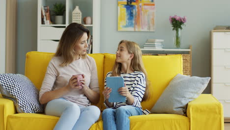 Blonde-Woman-Holding-A-Cup-Of-Coffee-And-Talking-With-Her-Teen-Daughter-Who-Is-Holding-A-Tablet-1