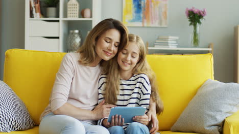 Blonde-Mother-And-Daughter-Resting-On-The-Sofa-At-Home-And-Watching-Something-On-Tablet,-Then-The-Mother-Hugs-Her-Daughter