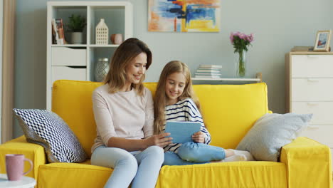 Blonde-Mother-And-Daughter-Smiling-And-Watching-Something-On-Tablet-While-Sitting-On-Sofa-In-Living-Room