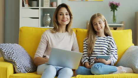 Blonde-Mutter-Und-Tochter-Sitzen-Auf-Einer-Gelben-Couch,-Während-Sie-Laptop-Und-Tablet-Benutzen-Und-Im-Wohnzimmer-In-Die-Kamera-Schauen
