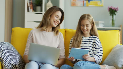 Blonde-Mother-And-Daughter-Sitting-On-Yellow-Couch-While-They-Using-Laptop-Computer-And-Tablet-And-Talking-In-The-Living-Room