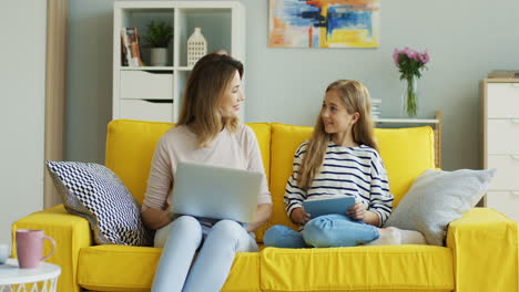 Blonde-Mother-And-Daughter-Sitting-On-Yellow-Couch-While-They-Using-Laptop-Computer-And-Tablet-And-Talking-In-The-Living-Room-1