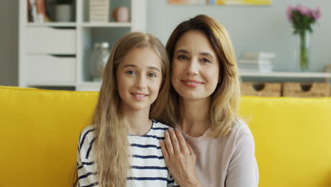 Portrait-Of-Blonde-Mother-And-Teen-Daughter-Sitting-On-Yellow-Sofa-And-Looking-At-Camera-While-They-Smile