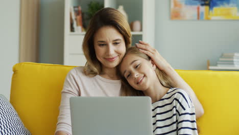 Cute-Teenage-Girl-Leaning-On-Her-Mother's-Shoulder-While-Watching-Something-On-Laptop-In-Living-Room