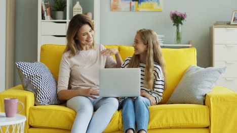 Blonde-Woman-Showing-Something-To-Her-Teenage-Daughter-On-Laptop-And-Talking-With-Her-While-They-Are-Sitting-In-Living-Room